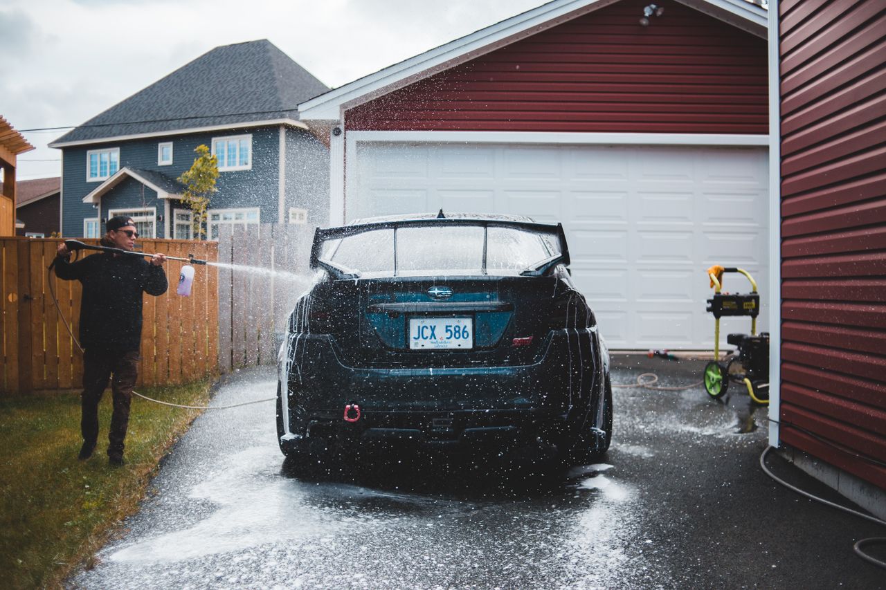 Full body of young male in black wear using long thin hosepipe while washing luxury vehicle in yard of tall private house